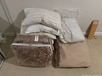 Lot of bedding items including comforters, pillows, mattress pad, as seen stacked on a floor.