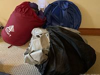 Camping lot with trekking sleeping bag in red carrying bag, blue collapsible basket, and black deflated Coleman queen size double high air mattress.