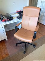 Front angle view of the Ikea Malcolm office chair on casters positioned on the clear plastic chair mat on hardwood floor, showing tan artificial leather upholstery and black armrests and base.