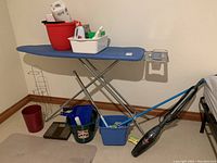 Side view of ironing board with buckets and cleaning supplies on and around it including mop and handheld vacuum