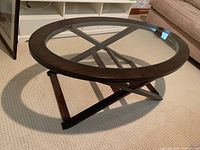 Oval coffee table with a dark wood frame and clear glass top, showing crossed wood support and wooden legs on carpeted floor near furniture.
