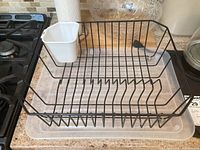 Black metal dish rack with white plastic utensil holder sitting on clear plastic tray on countertop near stove and paper towel roll.