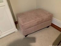 View of the pinkish ultra suede ottoman against two walls showing upholstery and cushion top