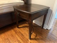 Side view of the dark wood side table next to a bed showing the curved edge, legs, and part of the attached power cord.