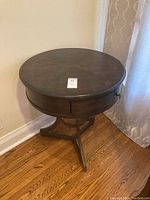 Full view of the wooden round side table showing the tabletop surface and tripod base, placed on hardwood floor near a curtain.