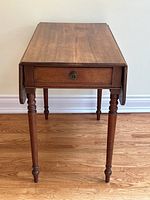 Mahogany drop leaf table showing full front view with drawer and leaves down, set on hardwood floor against wall.