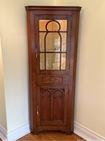 Full front view of walnut corner cabinet showing glass door, drawer, and lower panel.