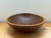 Front perspective view of the antique hand made wooden bowl on a wooden surface against a plain wall.