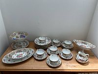 Full view of all cups, saucers, plates, platters, baskets and compote on wooden table against white wall.