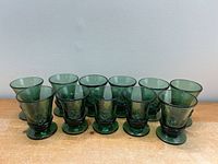 Front view of all eleven dark green hand-blown glasses arranged in two rows on wood surface against white wall.