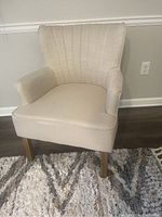 Front angle of beige upholstered accent chair showing vertical channel tufting on backrest and wooden legs on a area rug.