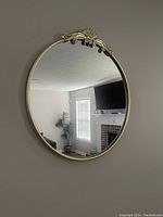 Front view of a round wall mirror with a gold frame and decorative floral and scroll design at the top center, reflecting a living room with window blinds, a TV, and a plant.