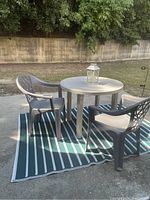 Wide shot of gray plastic round patio table and two chairs on green and white striped rug outdoors with trees and concrete wall in background.