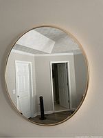 Photo shows a round mirror hanging on a light-colored wall reflecting a room with doors and ceiling details. Mirror has a thin gold metallic frame that is smooth and shiny.