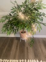 Artificial leafy tree in a woven basket-style pot on metal stand with string lights turned on.