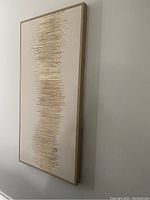 Side angled view of rectangular wall art with gold rods arranged vertically in a wavy pattern on beige canvas.