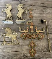 Top-down view of all six brass items on a wooden surface: two unicorn bookends, brass unicorn key hanger, enamel-on-brass hanging candleholder, and brass candle snuff stick.
