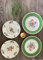Top down image of 4 floral china plates and one pressed green glass plate on wooden floor.