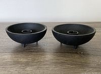 Pair of black cast iron artisan made candleholders shown side by side on a wooden surface under neutral lighting.