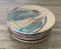 Stack of six pottery plates showing the top plate's abstract pattern with teal, black, and beige glazes on a wooden surface.