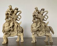 Front view of both faux ivory Asian sculptures side by side showing robed figures with children riding stylized lions.