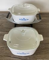 Two square CorningWare casserole dishes with glass lids. The larger 10 cup dish is seated in metal warming tray with wooden handles, all on wooden tabletop.