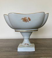 Full side view of the white porcelain footed fruit bowl showing gold floral designs and gold trim on the scalloped edges, pedestal, and base.