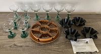 Eight handblown margarita glasses with green stems, round woven pottery serving dish with compartments, 4 black flower-shaped pottery tortilla pans, and some packaging paper.