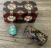 Top view of rosewood jewelry box with mother of pearl circular inlays and brass metal corner accents alongside jade egg on wooden stand and black-gold fabric case for opera glasses.