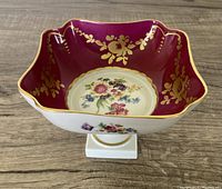Front angled view of square porcelain pedestal dish with burgundy, gold floral accents and central bouquet