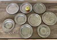 Overview of the nine antique pewter plates arranged on a wooden surface with an apple for size reference.