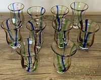 Ten artisan hand-blown drinking glasses arranged in a group, showing decorative vertical blue, green, and brown stripes on each clear glass.