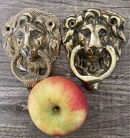 Two solid brass lion head door knockers side by side on wood surface, with an apple for size comparison.