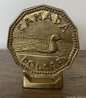 Front view of vintage brass coin bank resembling Canadian Loonie dollar with engraved loon and 'CANADA DOLLAR' text