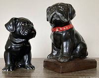 Front view of two black bulldog figurines, one smaller and smooth, the other larger with a red collar and wooden base.