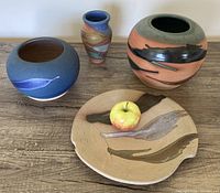 Four vintage signed art pottery pieces including three vases and one plate on a wood surface with an apple for scale.