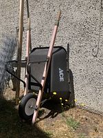 Full view of the wheelbarrow leaning upright against a cement wall outdoors on grass with some weeds.