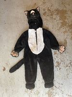 Full view of a black and white cat costume for children lying flat on the ground. Features hood with cat ears, front zipper, attached paws, and tail.