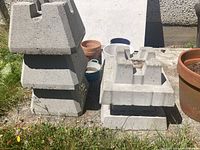 Photo shows cement blocks stacked in two piles on the grass next to a concrete wall. Some plant pots are visible in the background.