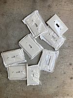 Eight white standard single toggle electric plates, new and still individually wrapped in clear plastic, laid out on a wooden surface.