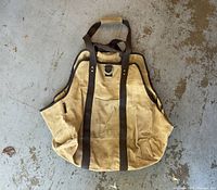 Full view of the beige canvas firewood carrier with brown trim and wooden handle placed on a concrete floor.