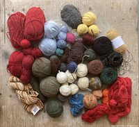 Photo of multiple yarn skeins and pompom balls spread out on wooden surface showing variety of colors and textures.