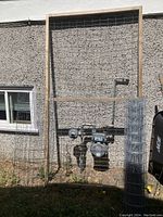 Large wooden frame with metal mesh attached standing outside against a textured wall, two metal gas meters just behind it.