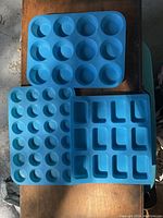 Three blue silicone baking pans on wooden surface: one 12-cup muffin pan, one 24-cup mini muffin pan, and one 12-compartment mini loaf pan.