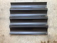 Top-down view of perforated steel baguette baking tray showing four curved channels with holes for airflow.