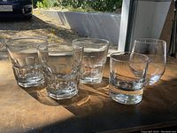 Photo of five whisky glasses on a wooden surface, illuminated by sunlight from the right. Three glasses have faceted and thick bases, the fourth is a lowball glass, and the fifth is a rounded stemless glass.