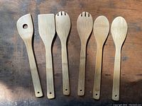 Photo showing six wooden serving utensils laid out on a wooden surface, including spatulas, forks, and spoons, all with holes in handles.