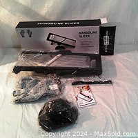 Photo showing mandoline slicer with box, cut-resistant gloves in plastic packaging, vegetable holder, and cleaning brush on white surface.