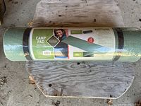 Green Wenzel Camp Pad rolled and sealed in packaging, placed on rustic wooden stool, showing label with size and brand.