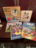 A photo showing 7 children's books in German including "Wieviel Uhr ist es, Lieber Bär?" with a clock illustration, "Guten Abend, gute Nacht!", "Der Maulwurf rettet das Häschen", "Tischlein Deck Dich", and "KNAX" magazine.